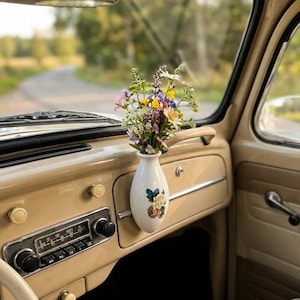 May include: Inside a classic car, a small white vase with a floral pattern holds a vibrant bouquet of wildflowers. The beige dashboard includes a radio. The car is parked on a road, with a view of trees.