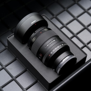 May include: A black Sony FE 1.4/35 GM camera lens with a lens hood, resting on a black box. The lens has a ribbed focus ring and various markings. The background is a dark, textured surface.