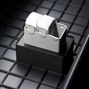 May include: A gray DJI drone battery charging case with two batteries, resting on a black box. The box sits on a black, grid-patterned surface. The DJI logo is visible on the case.