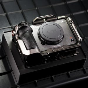 May include: A silver and black camera with a black grip and lens cap, resting on a black box. The lens cap has the Sony logo. The camera is in a metal cage with the word "Full-Frame" printed on the front.