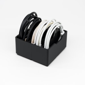 May include: A black rectangular organizer filled with neatly arranged charging cables. The cables are a mix of black and white, creating a contrast against the dark container. The organizer is designed to keep cables tidy.