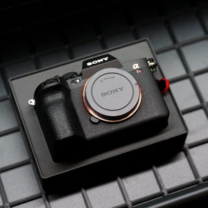 May include: A black Sony α7R camera with a lens cap, resting on a black box. The lens cap has the Sony logo. The camera has a textured grip and a red strap. The background is a dark, grid-like surface.
