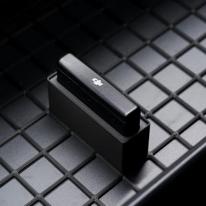 May include: A black rectangular device with a white logo rests on a black rectangular base. The base sits on a textured, grid-patterned surface. The image is well-lit, highlighting the device's sleek design and the texture of the surface.