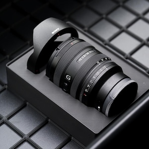 May include: A black Sony FE 2.8/16-35 GM camera lens with a lens hood, presented in a black box. The lens has a series of rings and markings, including the "G" logo. The background is a dark, textured surface.