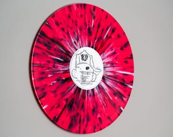 May include: A red and black splatter vinyl record with a white label featuring a cartoon character wearing a hat. The label text reads "The Electrocuted 80's" and lists the song titles.