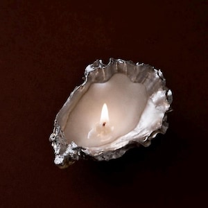 May include: A silver-toned oyster shell candle holder containing a white wax candle with a lit wick. The shell features a textured, irregular outer edge and a smooth, reflective interior surface that holds the melted wax pool around the flame.