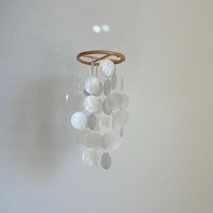 May include: A decorative wind chime featuring a natural woven ring at the top, from which hang multiple, iridescent, round, white shell discs. The discs are suspended by thin, clear strings, creating a shimmering effect.