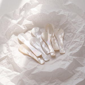 Puede incluir: Un conjunto de pequeñas cucharas nacaradas con un brillo lustroso. Las cucharas están dispuestas sobre una superficie de papel blanco texturizado. Las cucharas tienen un cuenco liso y curvado y un mango delgado, ideal para servir porciones pequeñas.