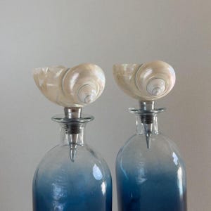 May include: Two glass bottles with blue ombre colouring, topped with seashell-shaped stoppers. The stoppers are a creamy white colour with spiral detailing. The bottles have silver-coloured metal accents.