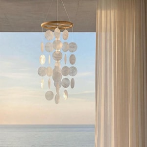 May include: A decorative wind chime with a woven circular top and numerous white, circular, and oval shell pieces hanging below. The chime hangs in front of a window overlooking the ocean.
