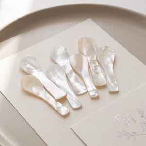 Puede incluir: Una colección de siete cucharas iridiscentes de nácar dispuestas sobre una superficie color crema. Las cucharas tienen un brillo nacarado y un cuenco pequeño y redondeado. Las cucharas están sobre papel color crema con un diseño floral.