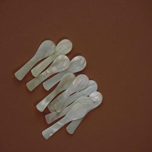May include: A collection of mother-of-pearl spoons, each with a pearlescent sheen, arranged on a brown surface. The spoons have a classic oval bowl shape and slender handles. The spoons are ideal for serving caviar or other delicacies.