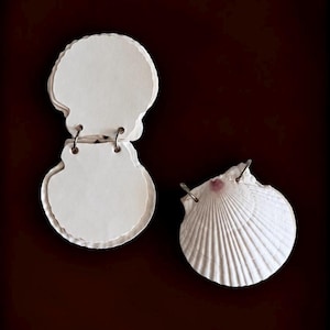Könnte beinhalten: Ein Set aus weißen, kammuschelförmigen Papierkarten, die mit silbernen Metallringen zusammengehalten werden, sowie eine einzelne weiße Kammuschel mit einem kleinen rosa Schimmer nahe dem Scharnier und silbernen Metallringen.