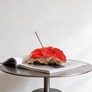 May include: A red and beige incense holder with a lit incense stick, emitting smoke. The holder sits on an open magazine on a metal table. The incense holder has a unique, organic shape and texture, adding a touch of natural elegance to the setting.