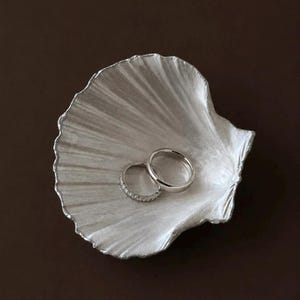 May include: A silver-toned, shell-shaped trinket dish with a textured, ribbed surface. Resting inside the dish are two rings: one plain, polished silver-toned band and one thinner silver-toned band set with a row of small, clear, sparkling stones.