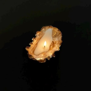 May include: A lit candle contained within a natural oyster shell. The interior of the shell features a metallic gold finish, while the wax is white and shaped to fit the irregular contours of the shell. The flame is bright and centered in the wax.