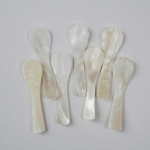 May include: A collection of seven small, pearlescent white spoons. Each spoon has a rounded bowl and a long, slender handle. The spoons are arranged on a plain white surface, showcasing their natural, iridescent sheen. Ideal for serving caviar or other delicacies.
