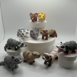May include: A collection of small, colorful animal figurines, including a tiger, raccoon, cat, ferret, opossum, and several mice. The figurines are arranged on a white tiered display stand.