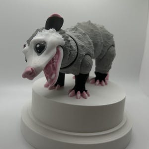 May include: A gray and white opossum figurine with pink ears and claws. The figurine is standing on a white pedestal.
