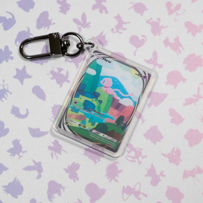 May include: A clear plastic keychain featuring a Pokemon card with a holographic effect. The card features an illustration of Mew, a pink mythical Pokemon, with the text "Mew" and "Pay Report" on the card.