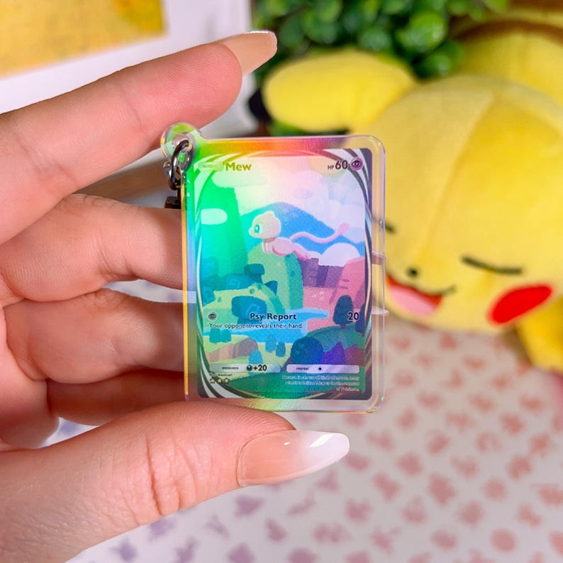 May include: A holographic Pokemon card keychain featuring Mew, a pink and white mythical Pokemon. The card has a rainbow iridescent finish and features the text "Mew" and "Psy Report".