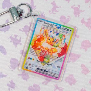May include: A clear plastic keychain with a colorful Eevee EX trading card. The card features a vibrant illustration of Eevee with rainbow accents and the text "Rainbow DNA" and "Coruscating Quartz". The keychain has a silver clasp.