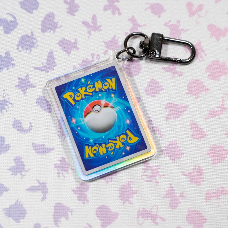 May include: A clear plastic keychain featuring a holographic Pokemon card with the Pokemon logo and a Pokeball. The keychain has a black metal clasp.
