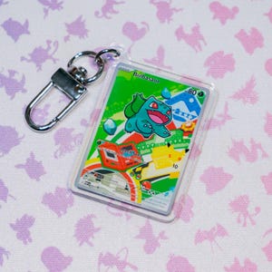 Op de afbeelding: Een doorzichtige acryl sleutelhanger met een rechthoekige ruilkaart met een groene Bulbasaur-karakterillustratie, de tekst Bulbasaur, Leech Seed en 80 HP, en een zilverkleurige metalen karabijnhaak bevestigd aan de bovenste hoek.