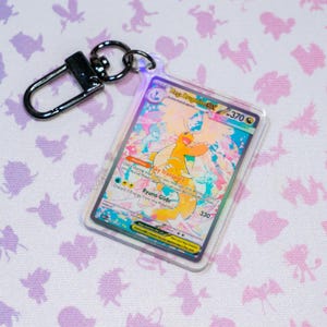 Op de afbeelding: Een doorzichtige acryl sleutelhanger met een Mega Dragonite GX ruilkaart met een kleurrijke, geïllustreerde draak. De kaart bevat de tekst Mega Dragonite GX, 370 HP, Sky Transport en Ryuno Glide. Inclusief een zwarte metalen karabijnhaak.