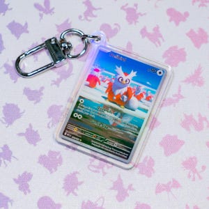 May include: A clear acrylic keychain featuring a Delibird trading card encased within a protective plastic sleeve. The card displays the character Delibird with the text Quick Gift and Gentle Slap. A silver metal lobster clasp is attached to the top.