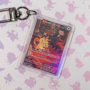 Op de afbeelding: Een doorzichtige plastic sleutelhanger met een Pokémon-ruilkaart met Meowth. De kaart heeft een kleurrijke illustratie van Meowth en een man in een pak. De kaarttekst bevat "Paw-cket Pilfer" en "Fury Swipes".