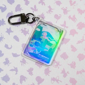 May include: A holographic keychain featuring the Pokemon Mew. The keychain is a clear plastic card with a holographic image of Mew and the text "Mew -60.0" and "Psy Report -20".
