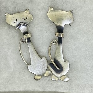 Puede incluir: Dos broches de plata en forma de gato con un diseño minimalista. Los gatos están mirando hacia adelante con los ojos cerrados y tienen una forma simple y redondeada.