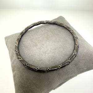 May include: A silver-toned bangle bracelet with a twisted rope design. The bracelet is circular and appears to be made of metal. The design features a series of intertwined lines that wrap around the bracelet. The bracelet is resting on a gray pillow.
