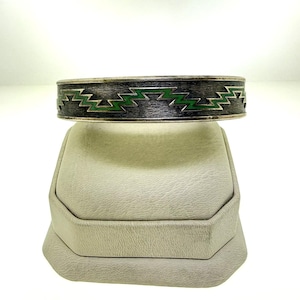 May include: A silver-toned cuff bracelet with a textured finish. The bracelet features a repeating zigzag pattern in green enamel. The design is set against a dark background, creating a striking contrast. This piece is a stylish accessory.