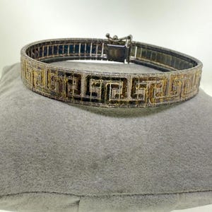 Könnte beinhalten: Ein metallisches Armband mit einem sich wiederholenden griechischen Mäandermuster in dunkler, oxidierter Optik. Das Band besteht aus verbundenen rechteckigen Segmenten und verfügt über einen sicheren Kastenschloss-Verschluss.