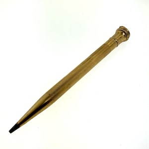 以下が含まれることがあります： 金色の仕上げが施されたヴィンテージのメカニカルペンシル。ペンシルはテクスチャード加工されたボディと装飾的なキャップを備えています。芯先が白い背景に映っています。