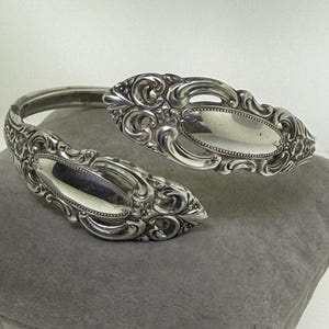 May include: A silver cuff bracelet with intricate floral patterns. The bracelet has two oval decorative sections with a reflective surface. The open design allows for easy wear. The bracelet is displayed on a grey cushion.