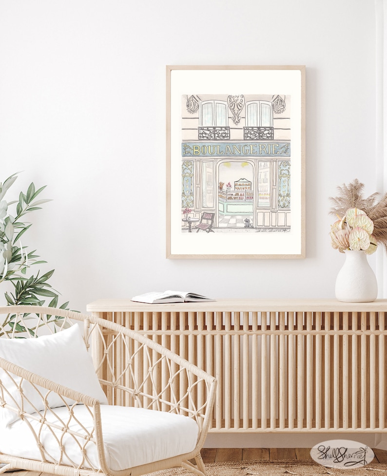 May include: A watercolor illustration of a Parisian bakery storefront with a blue sign that reads "BOULANGERIE". The bakery has a large window with a view of the interior, and there is a small table and chair outside.