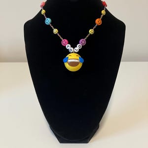 May include: A beaded necklace featuring a variety of colourful smiley face beads in shades of yellow, pink, orange, purple, and blue. The necklace includes white letter beads spelling out YOLO and a large yellow laughing emoji pendant.