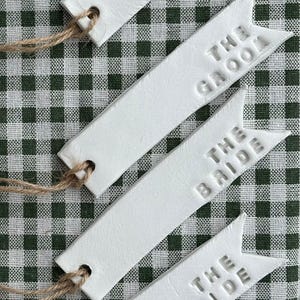 May include: White rectangular tags embossed with "THE GROOM" and "THE BRIDE". Each tag has a hole with natural twine. The tags are arranged on a green and white checkered background.
