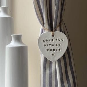 May include: White ceramic heart-shaped ornament with the text "Love you with my whole" and a small heart symbol. The ornament is hanging on a striped curtain.