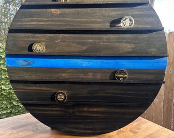 Op de afbeelding: Een ronde houten display met een blauwe streep in het midden. De display heeft vijf planken om kleine items op te bergen, zoals munten of badges. Er zijn vijf gouden badges op de planken, waaronder een met de tekst "TASER" erop.