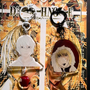 May include: Two acrylic keychains featuring anime characters. The left keychain has a white apple-shaped clasp, a white-haired character, and a red apple charm. The right keychain has a black apple-shaped clasp, a blonde character in a red hood, and a brown apple core charm.