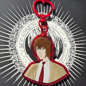 May include: An acrylic keychain featuring an illustration of a person with brown hair wearing a tan blazer, white shirt, and red tie. The keychain has a red apple-shaped clasp and a red border, set against a circular black and white design.