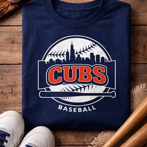 Puede incluir: Camiseta azul marino con un logotipo de béisbol de los Cubs en blanco y rojo. El logotipo presenta la palabra "CUBS" en rojo, un bate de béisbol y una silueta del horizonte. También se ven una pelota de béisbol, un guante y zapatillas.