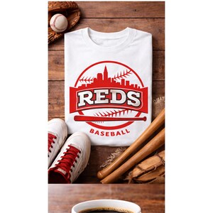 May include: White t-shirt with a red baseball-themed graphic featuring the word "REDS" and a cityscape design. The image includes a baseball, glove, bat, and white sneakers with red laces, all arranged on a wooden surface.
