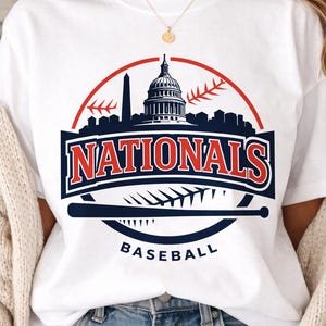 May include: White t-shirt featuring a baseball-themed graphic. The design includes the text "NATIONALS" in red and blue, a baseball bat, and a cityscape silhouette. The word "BASEBALL" is printed below the bat.