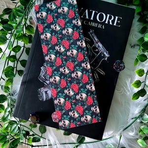 Puede incluir: Un marcapáginas decorativo con un patrón de calaveras blancas y rosas rojas sobre un fondo oscuro. El marcapáginas está colocado sobre un libro titulado "Salvatore Cabrera". La imagen también incluye un anillo y un broche en forma de rosa.