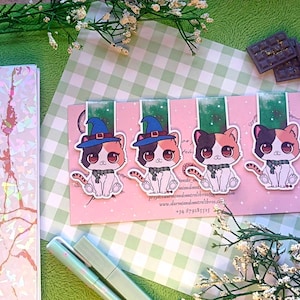 May include: Four cat-themed bookmarks with witch hats and scarves on a pink background. The cats are calico with large eyes. A notebook and pens are in the background. The bookmarks have the text "www.dormiendosentrelibros.com".